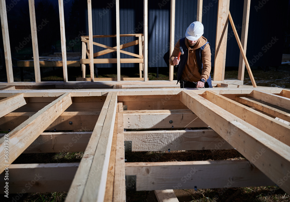 Man worker building wooden frame house on pile foundation. Carpenter hammering nail into wooden truss, using hammer. Carpentry concept. Man worker building wooden frame house on pile foundation. Carpenter hammering nail into wooden truss, using hammer. Carpentry concept.