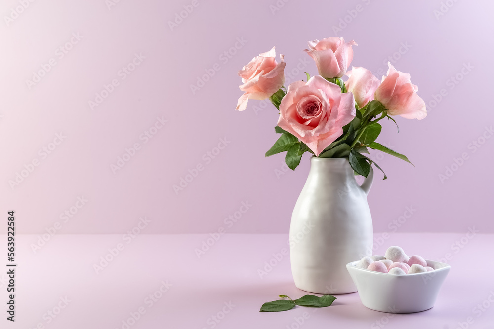 Pink roses in a white ceramic vase and round candies in a vase on a purple background. Card. Copy space. Photo.