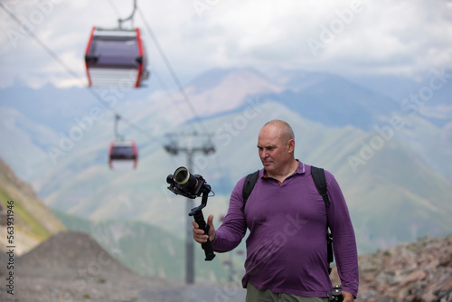 A man is filming a cable car in the mountains with a video camera. A tourist writes a YouTube blog while traveling.