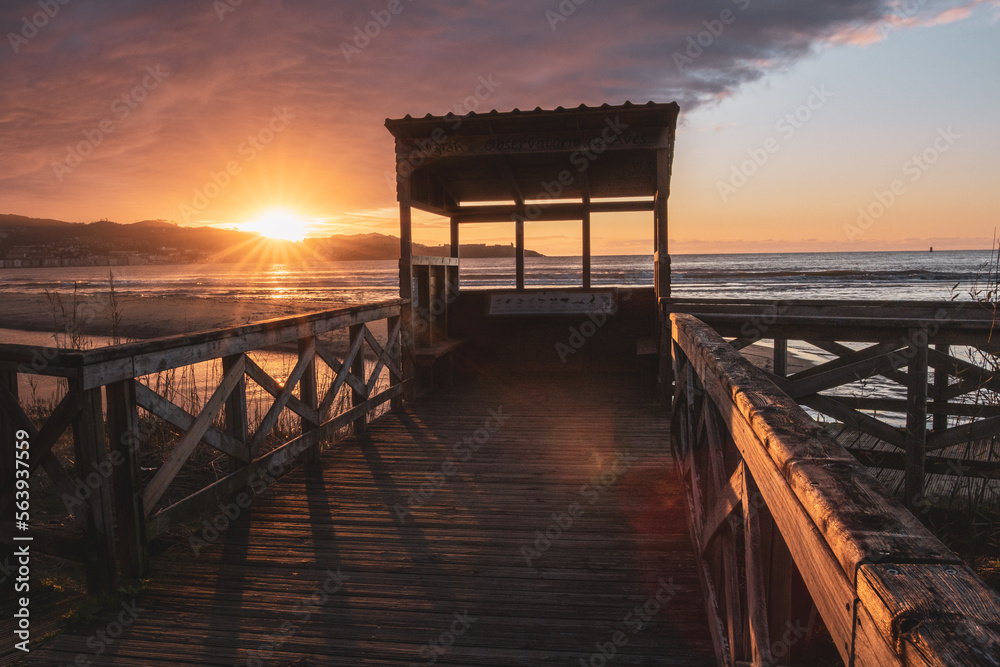 Bird hide on a wooden pier at sunset on Playa America beach. Sun star over the mountains on the coastline. Nigran, Pontevedra, Galicia, Spain. Bird hide on a wooden pier at sunset on Playa America beach. Sun star over the mountains on the coastline. Nigran, Pontevedra, Galicia, Spain.