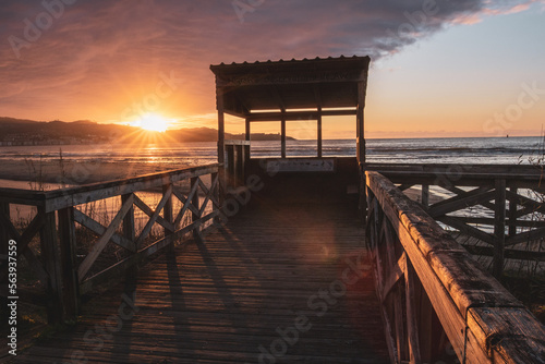 Bird hide on a wooden pier at sunset on Playa America beach. Sun star over the mountains on the coastline. Nigran, Pontevedra, Galicia, Spain.