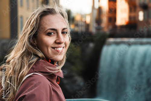 Beautiful girl near a large waterfall. Blonde tourist smiles looking at the camera. Young woman with windblown hair in Norrköping. Enjoyment of traveling and discovering the world. photo