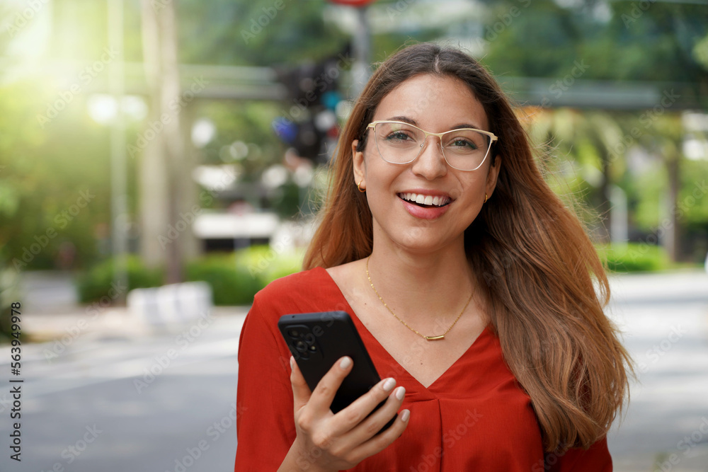 Brazilian woman looking at camera holding smartphone with blurred city street in Sao Paulo,  Brazil