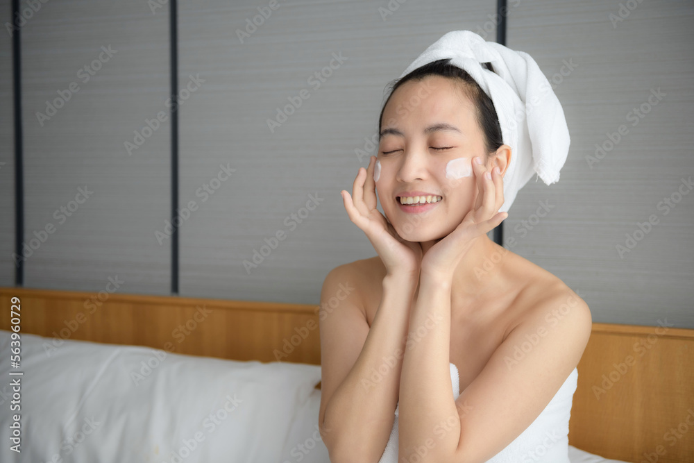 Happy young Asian woman applying face lotions while wearing a towel and touching her face. Daily makeup and skincare Happy young Asian woman applying face lotions while wearing a towel and touching her face. Daily makeup and skincare