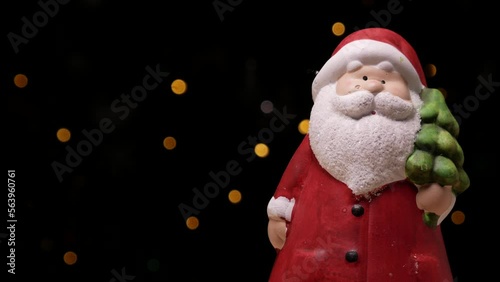 Ceramic Santa Claus figurine. Festive toy decoration for Christmas. Tree flickering lights bokeh in background. Xmas Holiday Season winter composition with copy space.