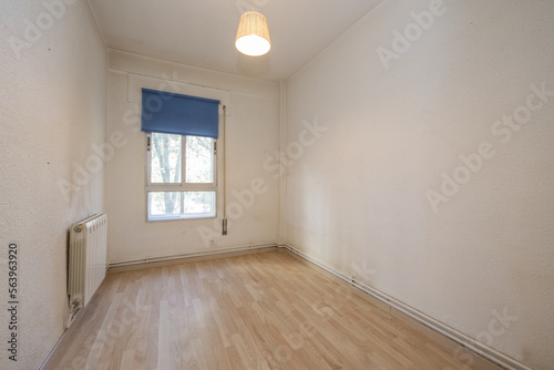 Old bedroom with a small aluminum window with a blue shutter, walls in need of a coat of paint and laminated flooring
