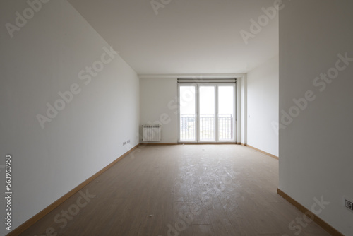 Empty room with light wooden floors  white aluminum radiators  plain white painted walls and a balcony with three aluminum and glass doors with a view