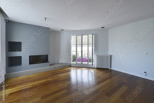 Empty living room with a dark oak hardwood floor  smooth light blue-grey walls  a built-in fireplace in the wall  and access to a terrace with awnings