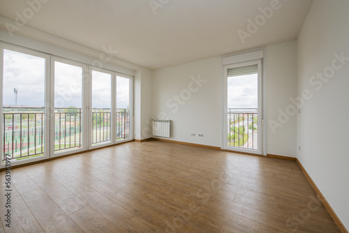 Empty room with light wooden floors  white aluminum radiators  smooth white painted walls and four aluminum and glass doors with views of the city and a second balcony on another wall