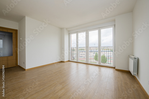 Empty room with light wooden floors, white aluminum radiators, smooth white painted walls, oak and glass front door and blacond e four aluminum and glass doors with views of the city