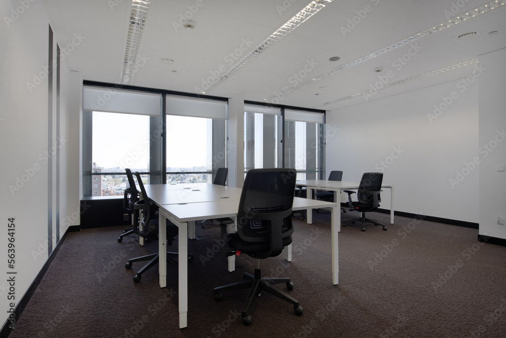 Spacious empty office with brown carpet and white wooden tables with swivel black seats and technical ceiling and large windows with views Spacious empty office with brown carpet and white wooden tables with swivel black seats and technical ceiling and large windows with views