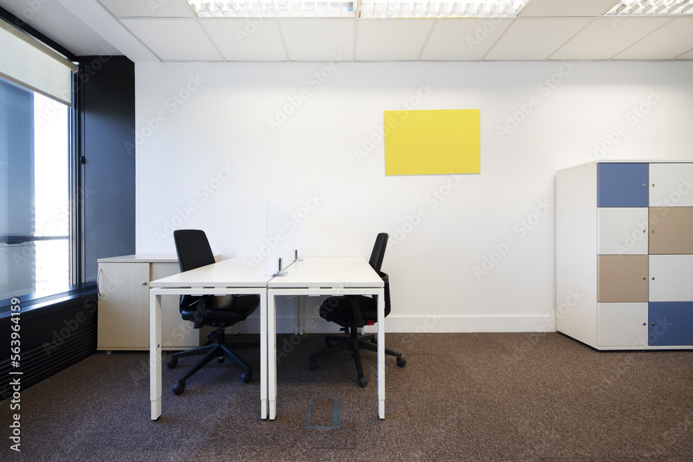 Spacious office with brown carpet, white wooden tables, filing cabinet with colored doors and black swivel chairs and bay window with view Spacious office with brown carpet, white wooden tables, filing cabinet with colored doors and black swivel chairs and bay window with view