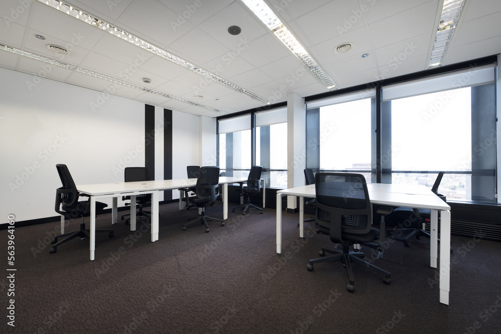 Spacious empty office with brown carpet and long white wooden tables with swivel black seats and technical ceiling and large windows with views Spacious empty office with brown carpet and long white wooden tables with swivel black seats and technical ceiling and large windows with views