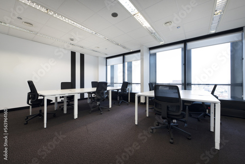 Spacious empty office with brown carpet and long white wooden tables with swivel black seats and technical ceiling and large windows with views