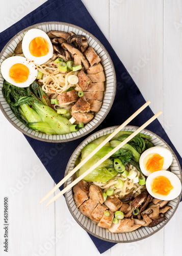 Two bowls of Asian noodle soup, ramen with chicken breast, vegetables, mushrooms, egg and chopsticks on a dark napkin, on a white wooden table. Top view, healthy eating concept