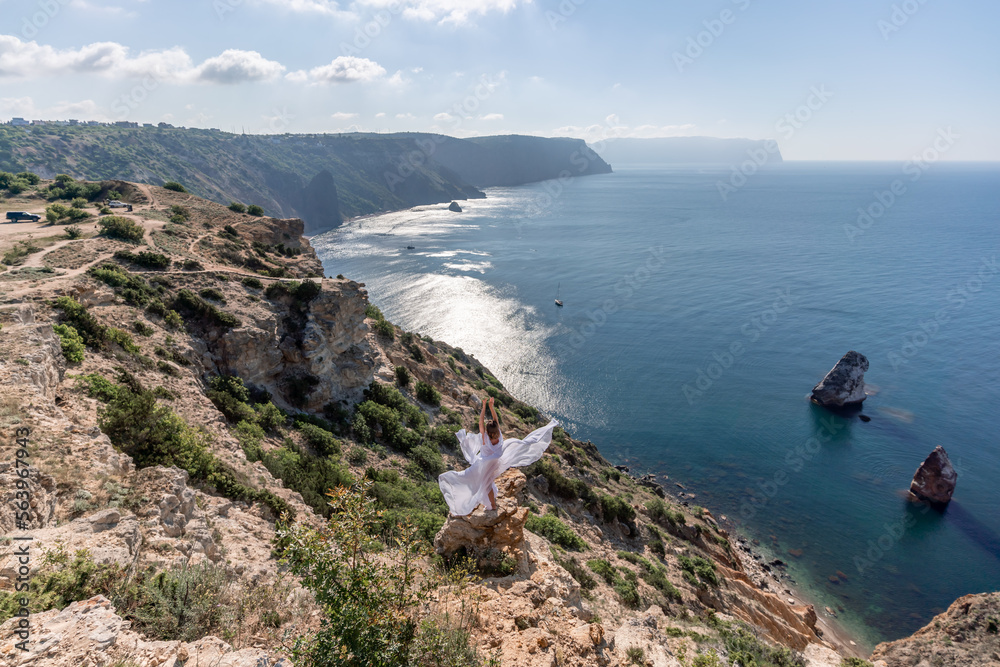 A beautiful young woman in a white light dress with long legs stands on the edge of a cliff above the sea waving a white long dress, against the background of the blue sky and the sea.