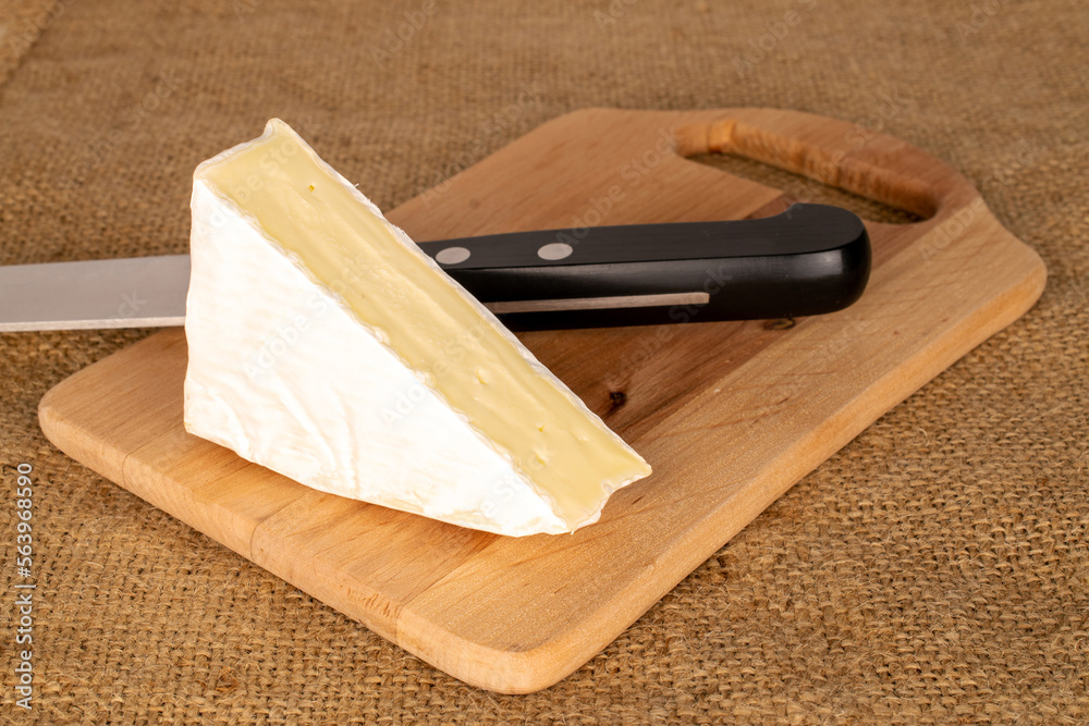One slice of delicious brie cheese with wooden kitchen board and metal knife on jute cloth, macro.