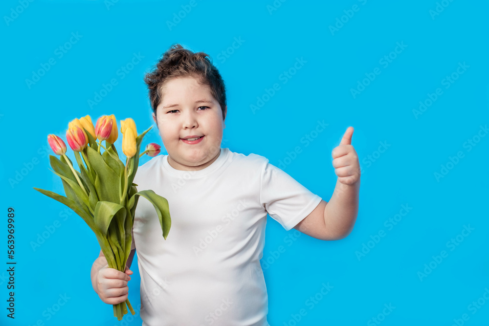 A cute little boy in a white T-shirt holds a bouquet of tulips and points his thumb up against a blue background