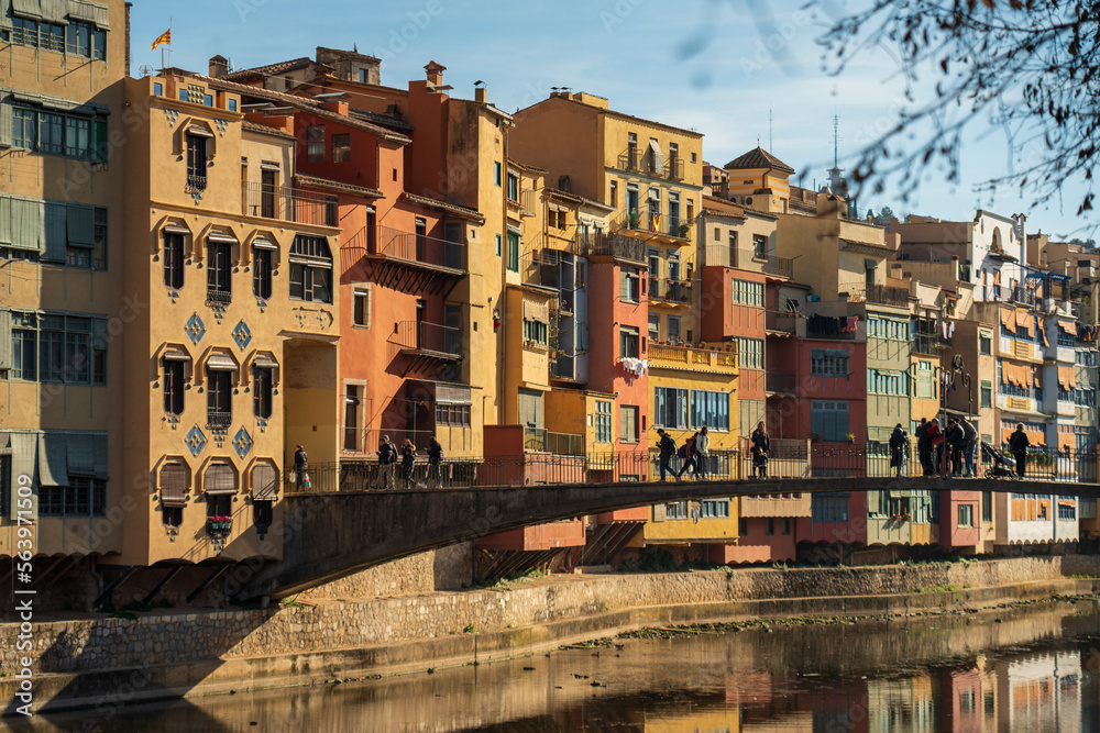 View of bright traditional Mediterranean houses on the canal's banks on a sunny day. People walk on the bridge in Girona city.