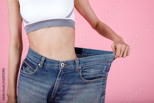 Woman shows weight loss. Slim woman is happy to show her big old jeans. Fit woman in oversize blue jeans on pastel pink background. Diet and weight loss concept.