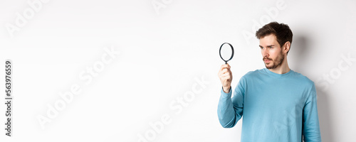 Young handsome man look through magnifying glass with curious face, investigating or searching for something, standing on white background