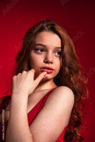 Pensive fashionable teen cover girl with curly hair in stylish red dress pose on red background, looking away. Young pretty brunette lady posing in studio. Fashion style concept. Copy space for ad