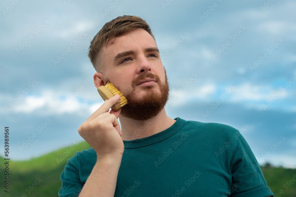 Handsome young bearded guy, European brutal man with a beard hair is combing the beard on face with a comb, brush. Male personal self care, products, accessory on nature. Natural summer background Handsome young bearded guy, European brutal man with a beard hair is combing the beard on face with a comb, brush. Male personal self care, products, accessory on nature. Natural summer background