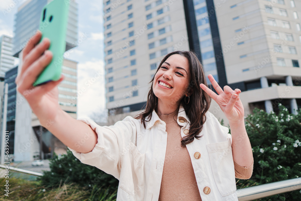 Fototapeta premium Close up portrait of one young woman smiling taking a selfie portrait with a cellphone, at background city buildings. Front view of a brunette happy girl doing a photo with a smartphone and sharing in