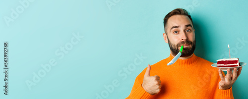 Close-up of funny adult man celebrating his birthday, holding bday cake with candle, blowing party wistle and showing thumb-up, standing over light blue background photo