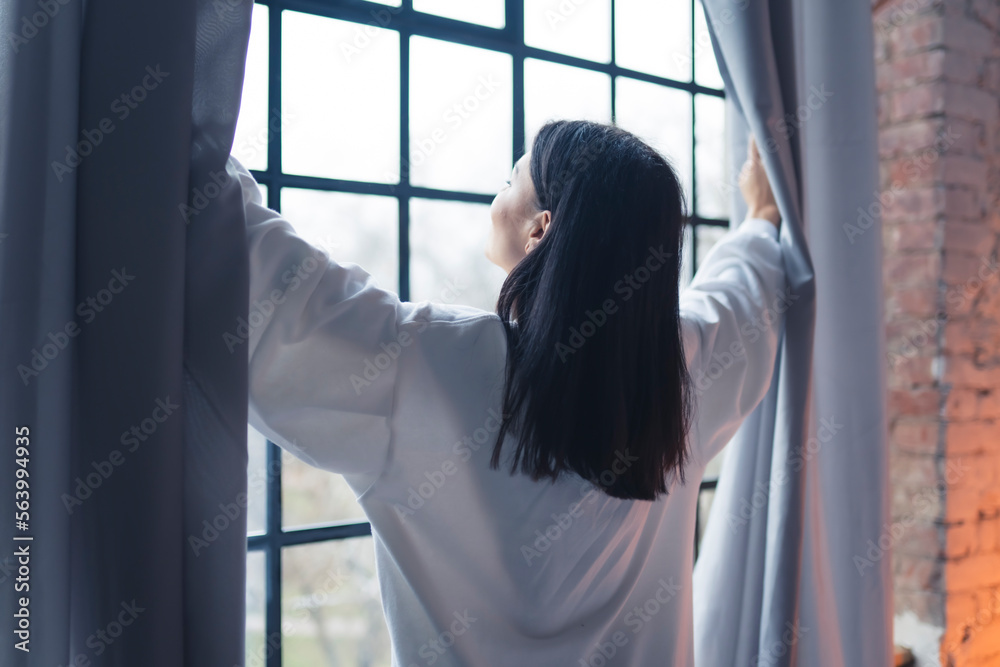 Back view of a woman opening window curtains and looking outside. No face. High quality photo