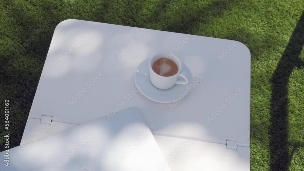 Taza de café con ordenador portátil en mesita de piscina sobre césped a la sombra de una palmera que se mueve por el viento.