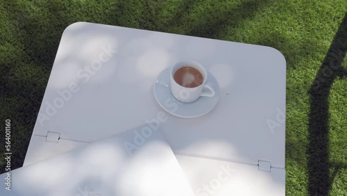 Taza de café con ordenador portátil en mesita de piscina sobre césped a la sombra de una palmera que se mueve por el viento. photo