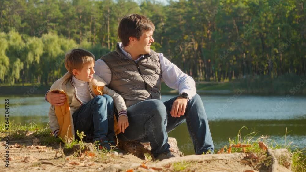 Slow motion of boy and father relaxing at riverbank while sitting on tree log. Family outdoors, happy parenting and childhood, autumn landscape, kids and parents relaxing outside Slow motion of boy and father relaxing at riverbank while sitting on tree log. Family outdoors, happy parenting and childhood, autumn landscape, kids and parents relaxing outside