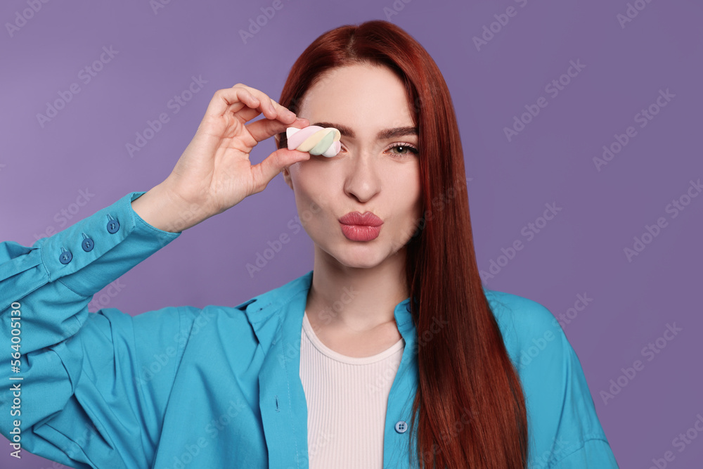 Obraz premium Beautiful woman with red dyed hair and marshmallow blowing kiss on purple background