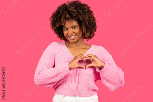 Happy pretty young african american curly woman with braces in casual outfit showing heart sign with hands