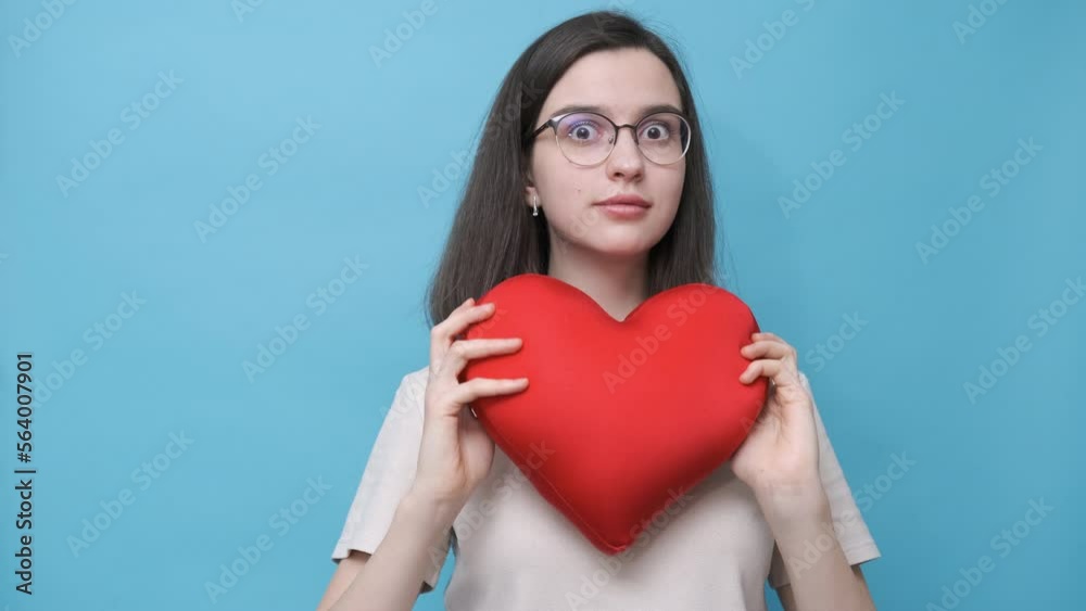 A beautiful student girl in glasses holding a red pillow in the shape of a heart and hiding and peeking out from behind it on a blue background. Love, relationships, Valentine's Day. A beautiful student girl in glasses holding a red pillow in the shape of a heart and hiding and peeking out from behind it on a blue background. Love, relationships, Valentine's Day.