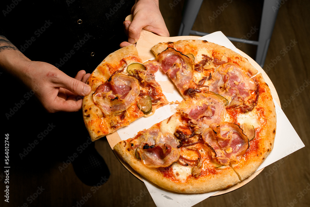 hand takes a piece of sliced tasty round pizza with bacon on wooden chopping board