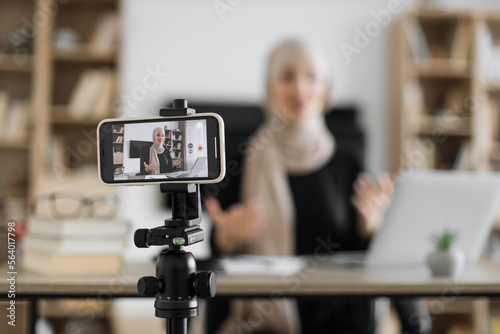Blur background of pretty muslim woman with headscarf sitting at desk and filming video blog. Focus on modern digital phone fixed on tripod.