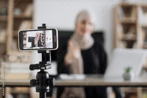 Focus on screen of phone of happy arab woman sitting at desk with books, notes and laptop talking with followers. Female tutor working at home during distance learning and showing thumb up.