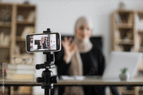 Focus on screen of phone of happy arab woman sitting at desk with books, notes and laptop talking with followers. Female tutor working at home during distance learning and showing sign ok.