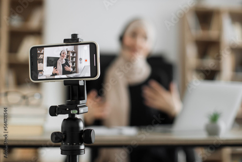 Charming muslim woman in hijab and headset talking and gesturing while recording video on modern phone. Female blogger sitting at home and doing live stream.