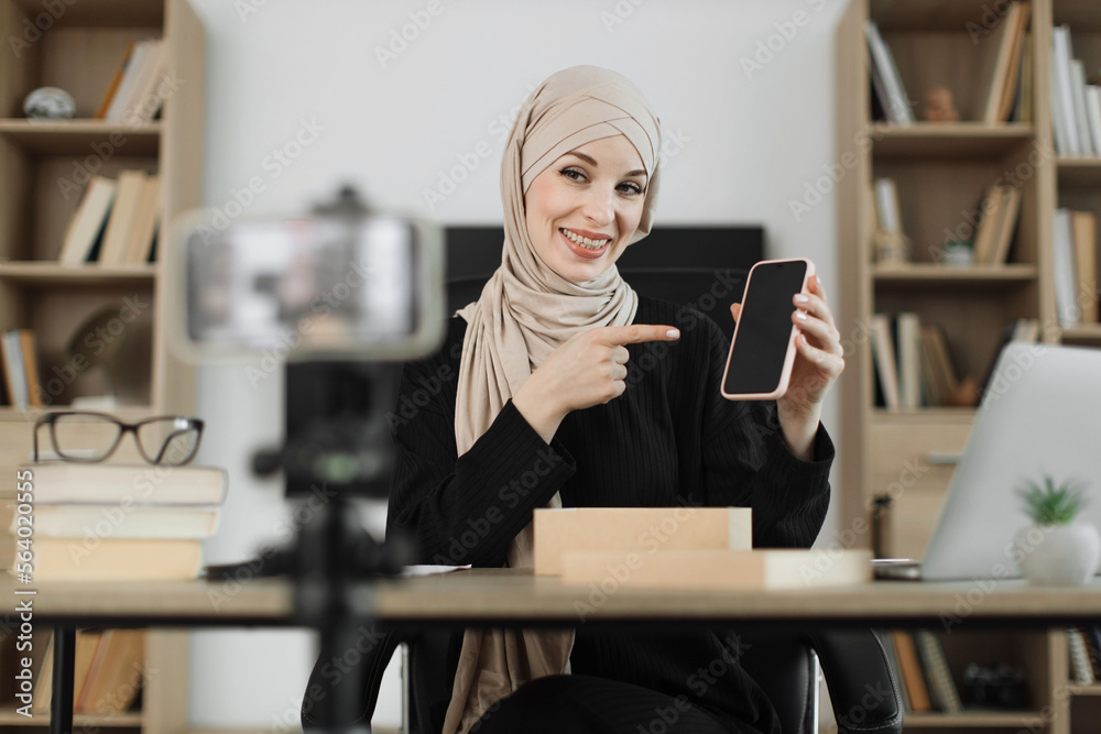 Pleasant muslim woman filming video on modern phone camera while opening parcel box with new smartphone. Concept of people, technology and blogging. Pleasant muslim woman filming video on modern phone camera while opening parcel box with new smartphone. Concept of people, technology and blogging.