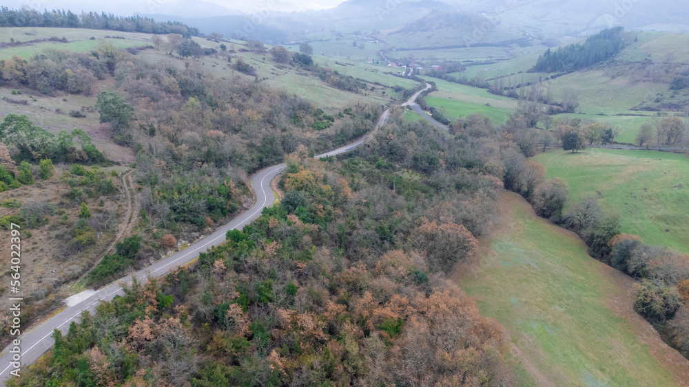 Aerial drone view of mountain road and green meadows on cloudy day with fog.