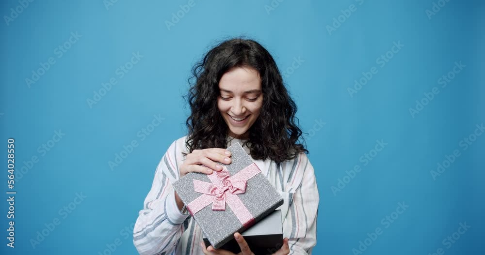 Cheerful brunette in striped shirt showing and tossing up silver gift box with with pink ribbon while looking at camera on blue background. Content female with shiny present box. Cheerful brunette in striped shirt showing and tossing up silver gift box with with pink ribbon while looking at camera on blue background. Content female with shiny present box.