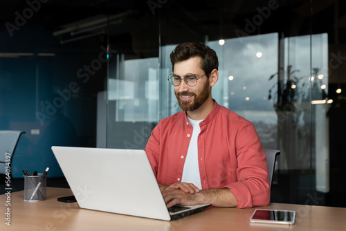 Fotografiet Successful smiling man working inside office with laptop, businessman in red shirt smiling and typing on keyboard in glasses, programmer working software for program