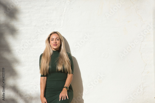 Young  pretty  blonde-haired  green-eyed woman in a green dress  leaning against a white wall  looking at the camera happy and smiling. Concept beauty  fashion  trend  eyes  happiness.