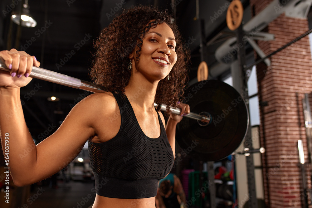 Fototapeta premium Sexy smiling gorgeous african american woman working out with barbell in gym