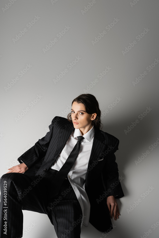 high angle view of brunette woman in black oversize suit and white shirt sitting and looking at camera on grey background. high angle view of brunette woman in black oversize suit and white shirt sitting and looking at camera on grey background.