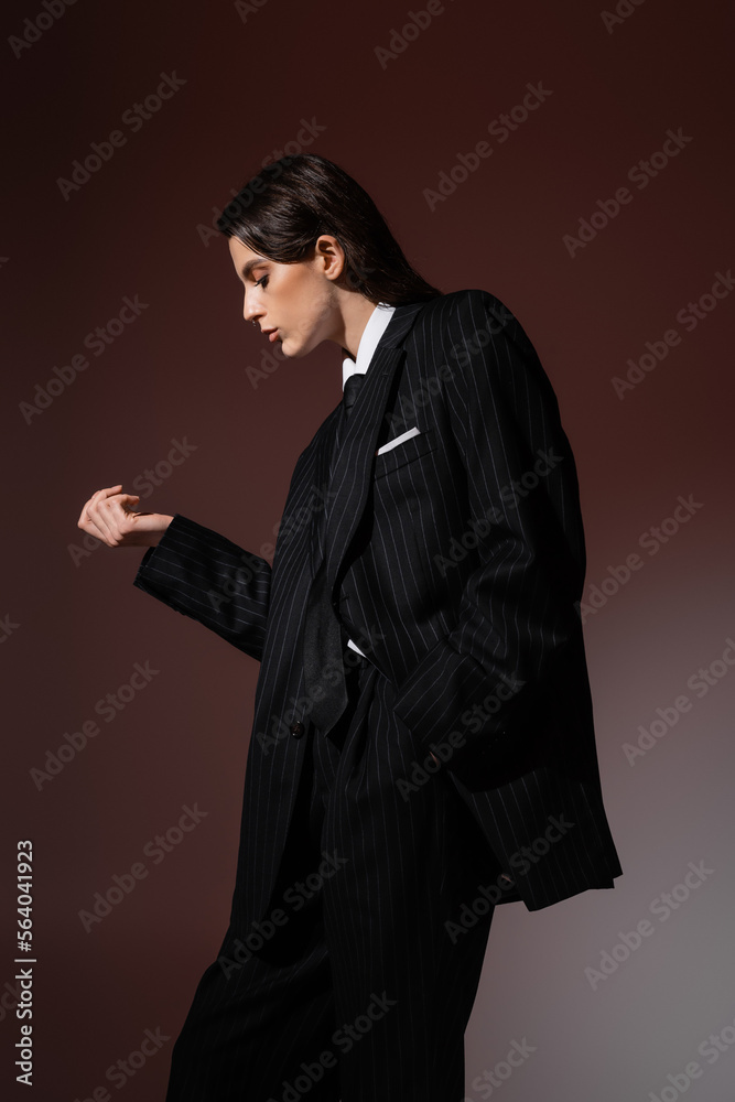 side view of brunette woman in black striped blazer posing with hand in pocket on brown background.