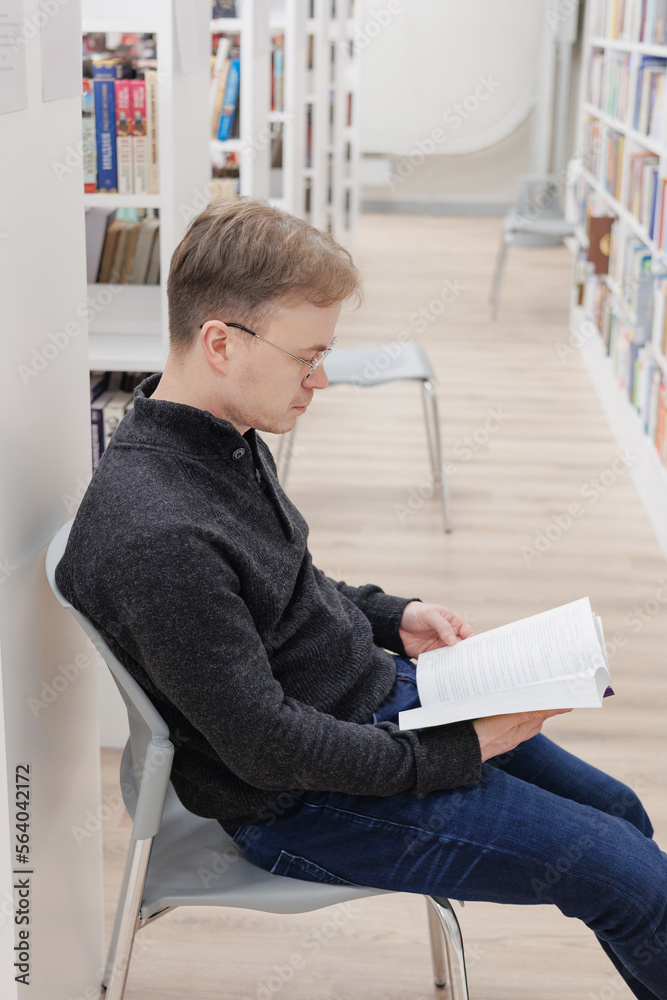 Obraz premium man in library is reading book sitting on chair near bookshelves. education writing scientific work, diploma. thoughtful young adult student or university teacher is learning foreign language